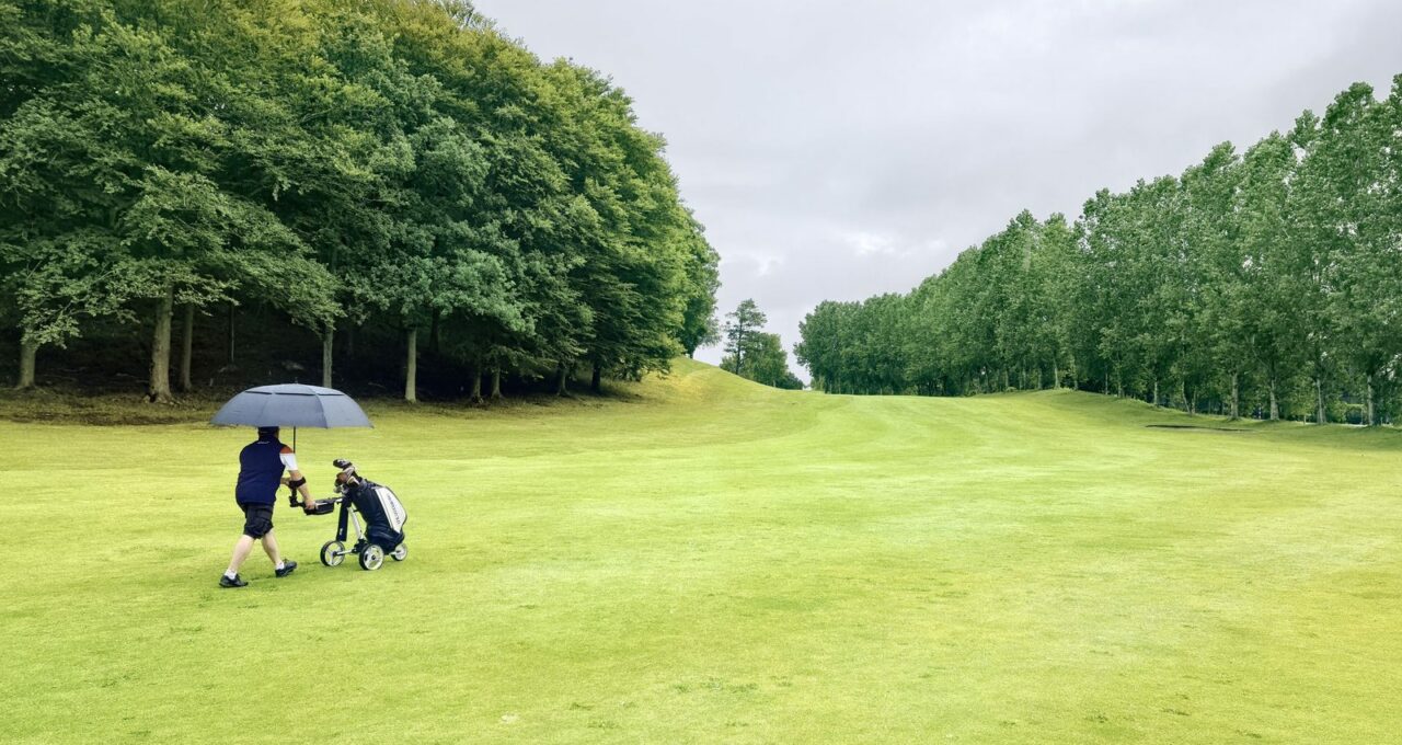 Deux joueurs traversent un fairway sous un parapluie, ciel bas.