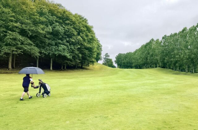 Deux joueurs traversent un fairway sous un parapluie, ciel bas.
