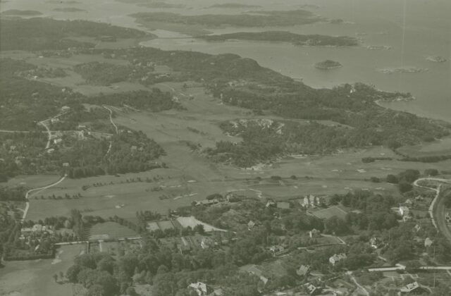 Golfbana med öppna spelfält och skog i bakgrunden.