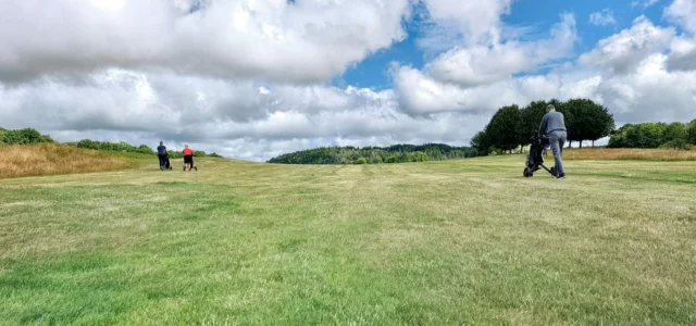 Three golfers walking up a fairway pushing their trolleys under a cloudy sky