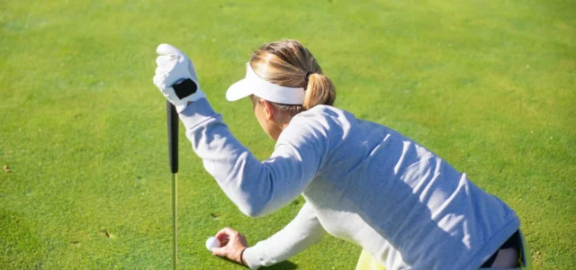 A golfer crouching on a green, leaning on her club, replacing her ball with bent knees