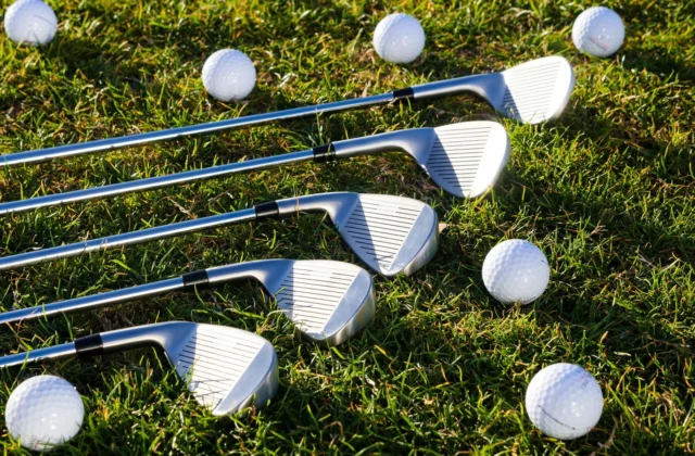 Five golf irons laid side by side on the grass, faces showing, surrounded by white golf balls