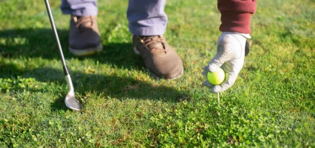 Hand in a white golf glove placing a yellow ball on a tee, a wedge resting on the grass beside it