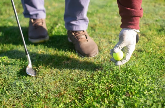Hand in a white golf glove placing a yellow ball on a tee, a wedge resting on the grass beside it
