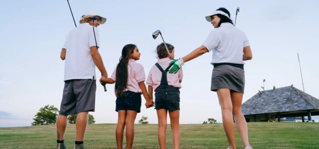 Two adults and two children seen from behind on a golf course, clubs in hand, the two girls holding hands