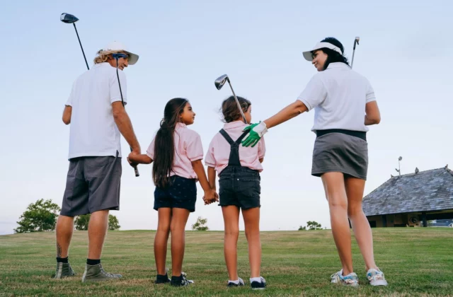 Zwei Erwachsene und zwei Kinder von hinten auf einem Golfplatz, Schläger in der Hand, die beiden Mädchen halten sich an der Hand