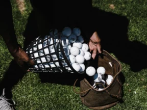 Hands tipping a wire basket full of used golf balls into a worn leather bag on the grass