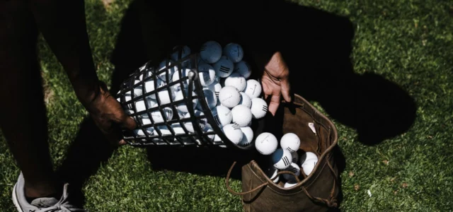 Hands tipping a wire basket full of used golf balls into a worn leather bag on the grass
