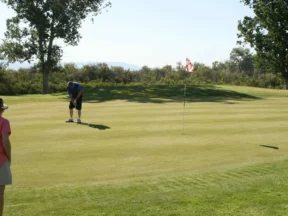 Three recreational golfers on a green: a man in blue strikes his putt while two women wait their turn, a red and white flag in the middle