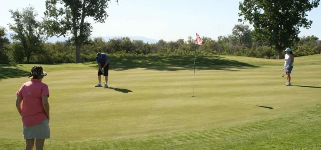 Drei Freizeitgolfer auf einem Grün: Ein Mann in Blau puttet, während zwei Spielerinnen warten, in der Mitte eine rot-weisse Fahne
