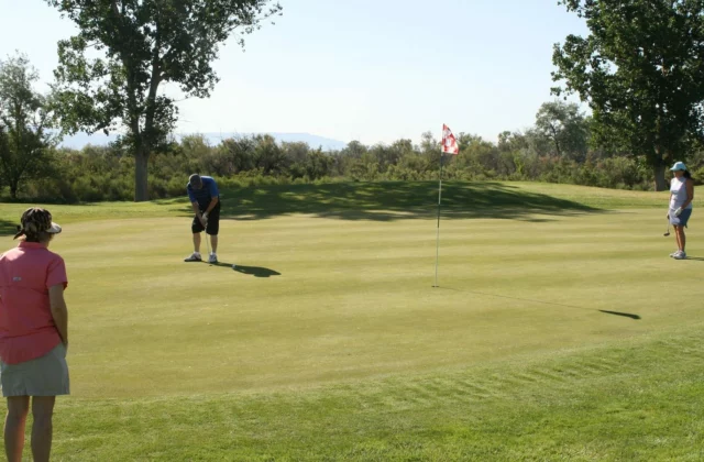 Drei Freizeitgolfer auf einem Grün: Ein Mann in Blau puttet, während zwei Spielerinnen warten, in der Mitte eine rot-weisse Fahne