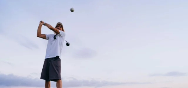 Un golfeur termine son élan au driver, la balle déjà en l'air, sous un ciel clair de fin de journée