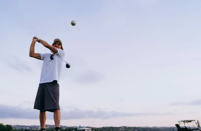 Ein Golfer beendet seinen Schwung mit dem Driver, der Ball fliegt schon, unter klarem Abendhimmel