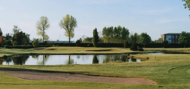 Un plan d'eau balisé de piquets rouges devant un green, avec un bunker au premier plan