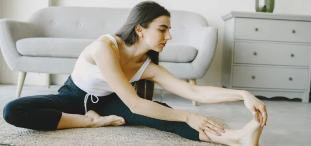 Une femme s'étire l'arrière de la jambe, assise sur un tapis dans son salon