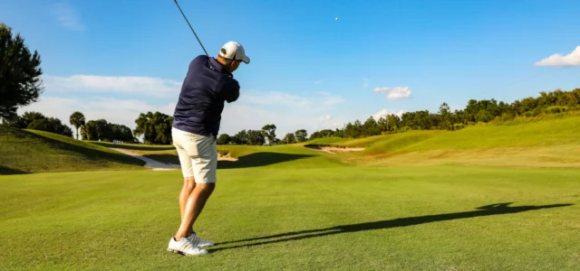 Un golfeur vu de dos termine un coup de fer sur un fairway ensoleillé, la balle haut dans le ciel bleu
