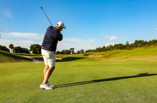 Ein Golfer, von hinten gesehen, beendet einen Eisenschlag auf einem sonnigen Fairway, der Ball hoch am blauen Himmel