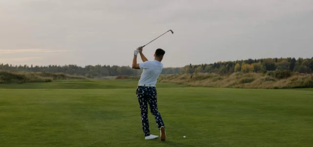 A golfer seen from behind, an iron raised above the shoulders, the ball on a fairway lined with autumn trees
