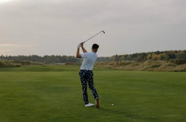 Golfer von hinten, das Eisen über die Schultern gehoben, der Ball auf einem Fairway mit herbstlichen Bäumen