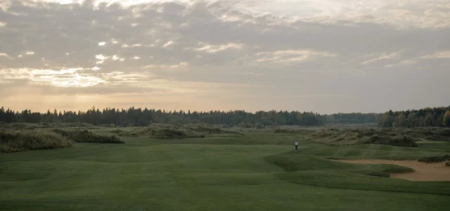 Un fairway vallonné sous un ciel voilé et une lumière basse, un golfeur seul au loin près d'un bunker