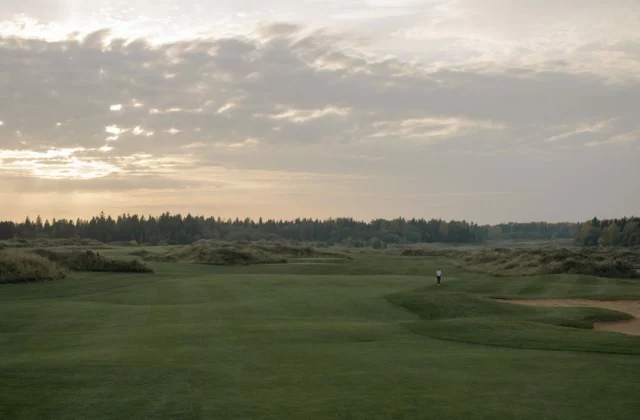 Ein welliges Fairway unter verhangenem Himmel und tiefem Licht, in der Ferne ein einzelner Golfer neben einem Bunker