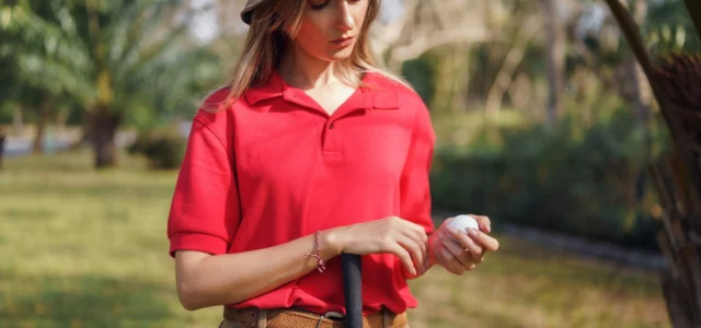 Golfeuse en chapeau à larges bords et polo rouge, une balle à la main, appuyée sur son club sous un soleil chaud