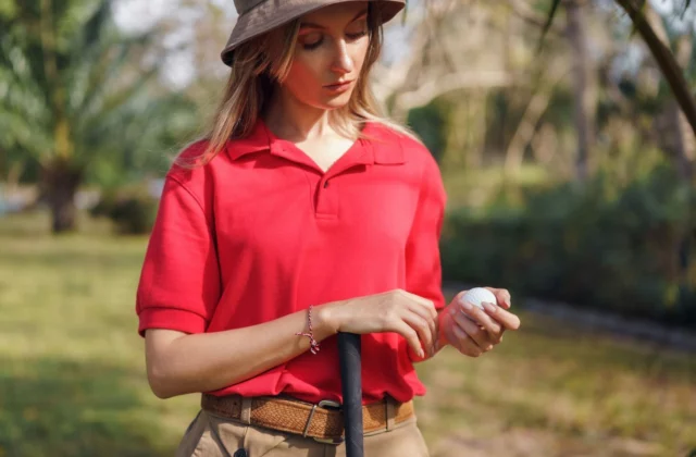 Golferin mit breitkrempigem Hut und rotem Poloshirt, einen Ball in der Hand, auf ihren Schläger gestützt in der heissen Sonne