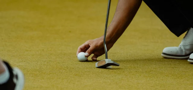 A golfer places a ball on the green just in front of a mallet putter held upright