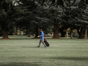 A golfer in a blue top, iron in hand, walking away from his stand bag on a fairway lined with tall conifers