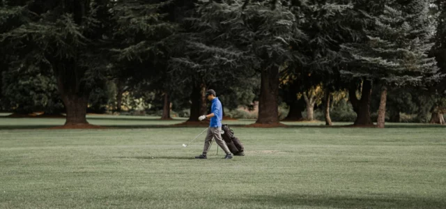 Golfeur en haut bleu, un fer à la main, qui s'éloigne de son sac posé sur ses pieds, sur un fairway bordé de grands conifères