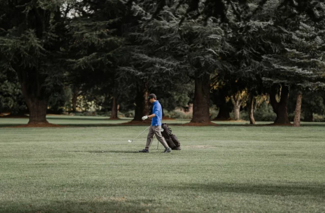 Ein Golfer in blauem Oberteil geht mit einem Eisen in der Hand von seinem aufgestellten Standbag weg, auf einem Fairway mit hohen Nadelbäumen