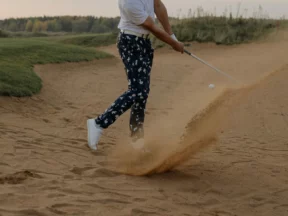 Un golfeur sort la balle d'un bunker au wedge, dans une gerbe de sable, en fin de journée