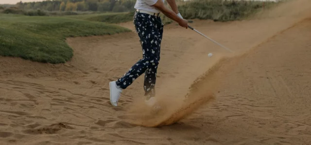 A golfer splashes the ball out of a bunker with a wedge in a spray of sand, late in the day