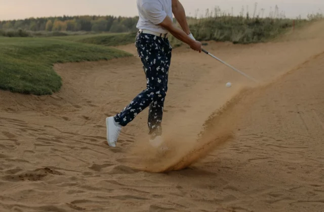 Ein Golfer schlägt den Ball mit dem Wedge aus dem Bunker, Sand spritzt auf, am späten Nachmittag